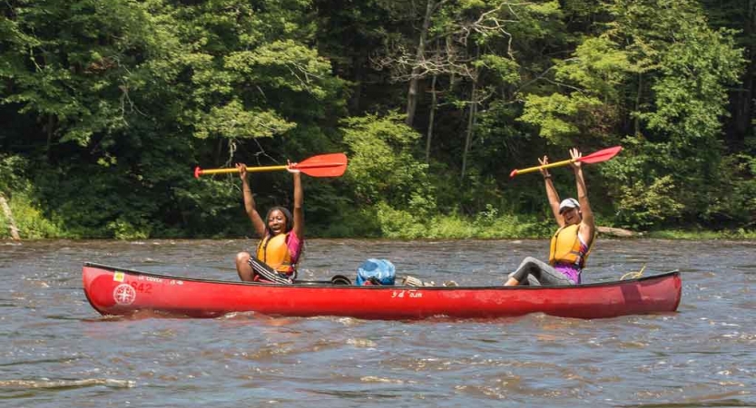 Two people wearing life jackets sit in a red canoe and hold their paddles in the air. 